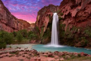 vibrant pink sky with red cliffs. and Havasu Falls waterfall flowing into a blue pool below.