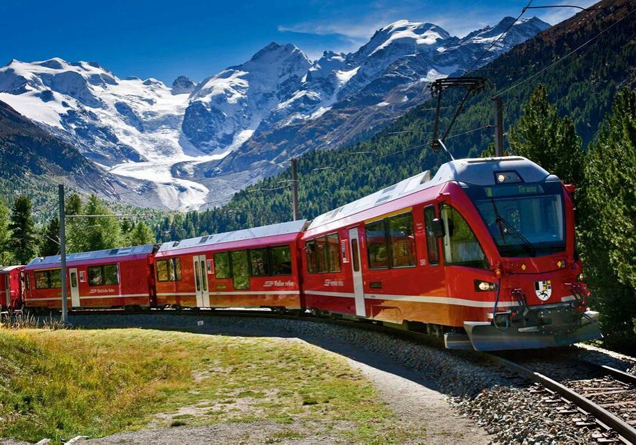 photo in Switzerlnd with the snow capped Alps in the background and a red passenger train in the foreground.